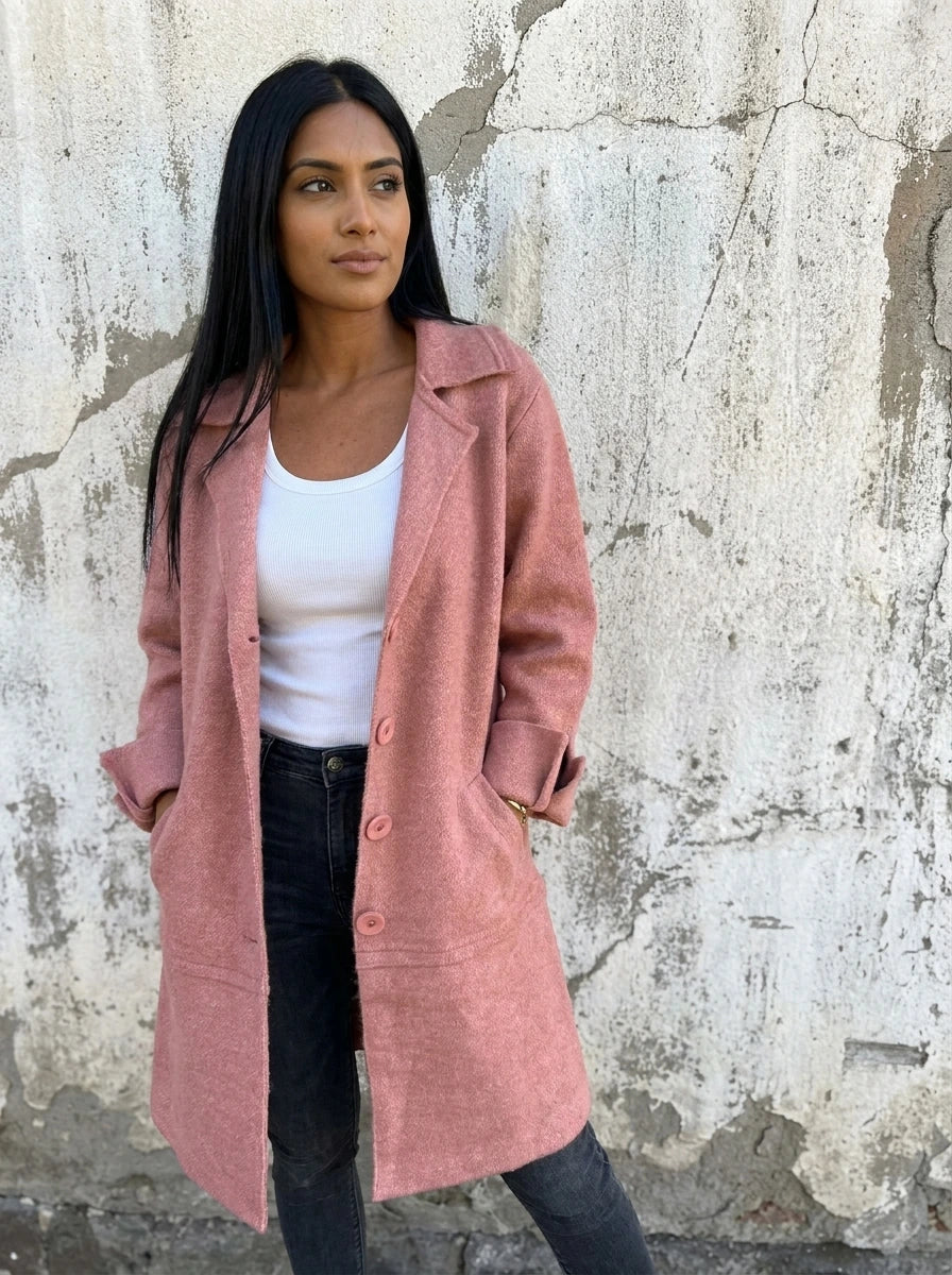 Woman wearing a dusty pink button-up long cardigan coat against a textured concrete wall