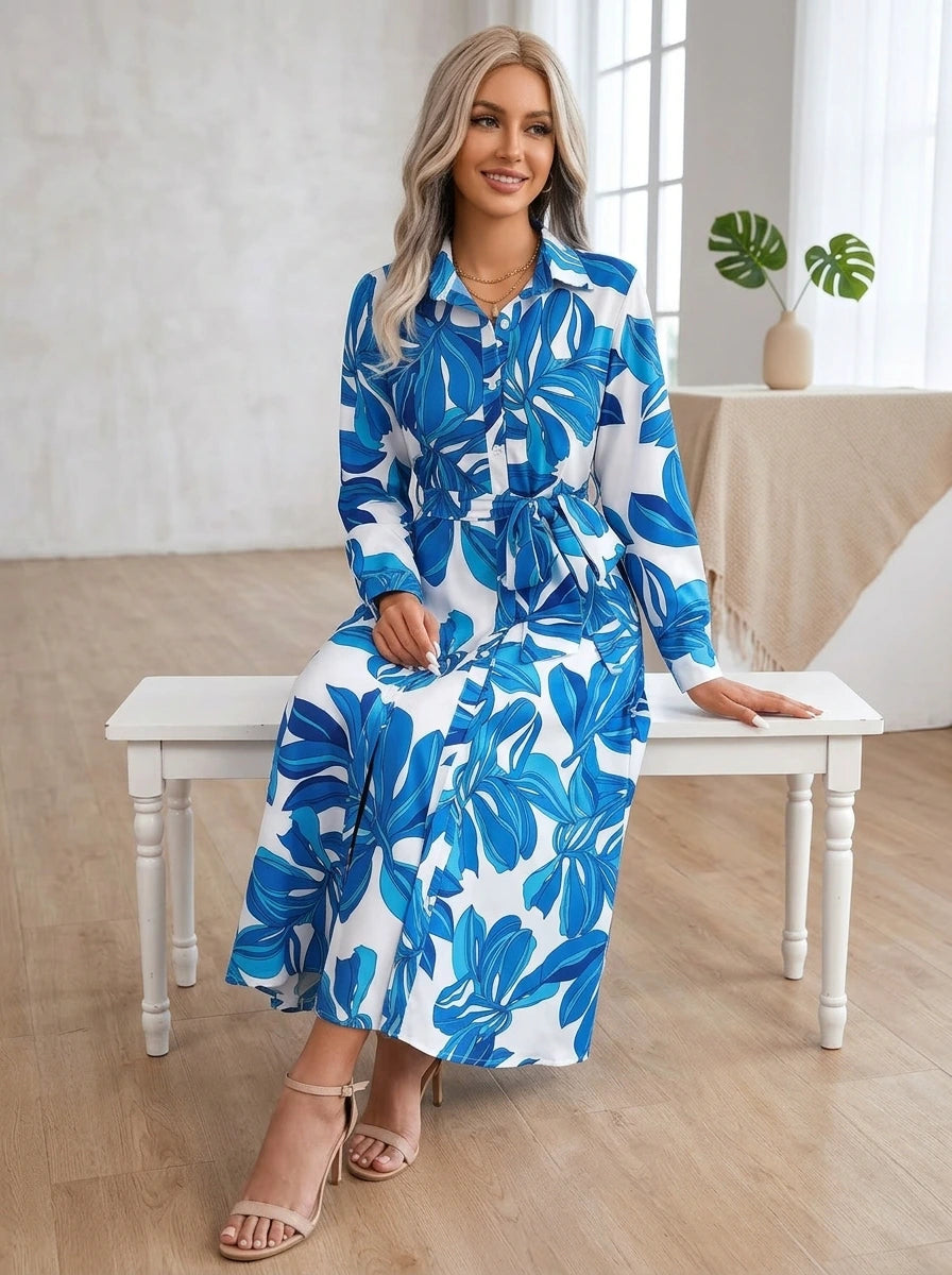 Driftly blue floral button-down midi dress with lapel collar, long sleeves, and adjustable waist tie, seated on a bench in a light, airy interior space.