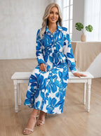 Driftly blue floral button-down midi dress with lapel collar, long sleeves, and adjustable waist tie, seated on a bench in a light, airy interior space.