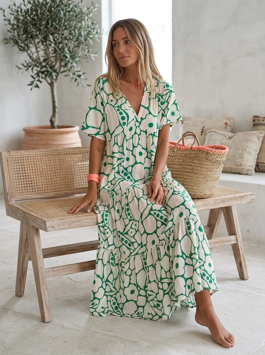 Driftly green floral V-neck midi dress, bohemian style, soft-blend fabric, seated on a bench in a light, airy interior space.