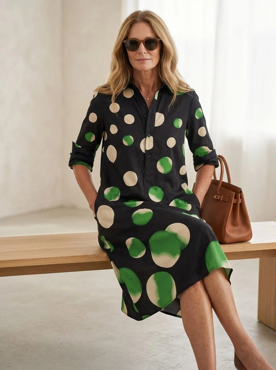 Driftly black polka dot collared midi dress with partial button front, three-quarter rolled sleeves, and side pockets, seated on a bench in a light, airy interior space.