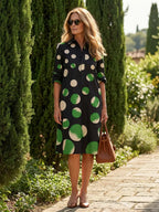 Woman in a black polka dot collared midi dress with green and cream spots, holding a brown handbag.