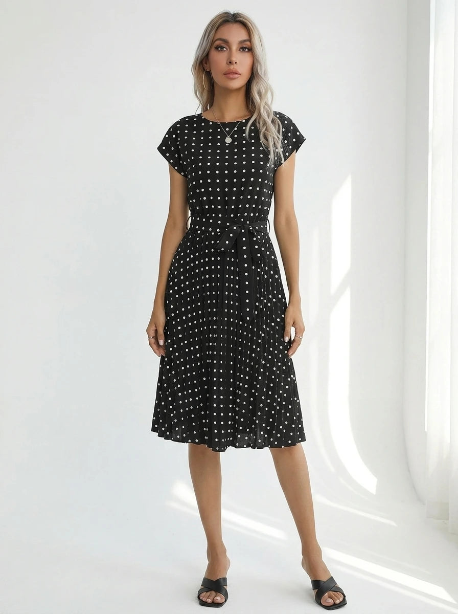 Driftly black polka dot pleated midi dress with cap sleeves and a self-tie waist belt, in a minimalist studio setting with soft window light.
