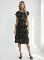 Driftly black polka dot pleated midi dress with cap sleeves and a self-tie waist belt, in a minimalist studio setting with soft window light.