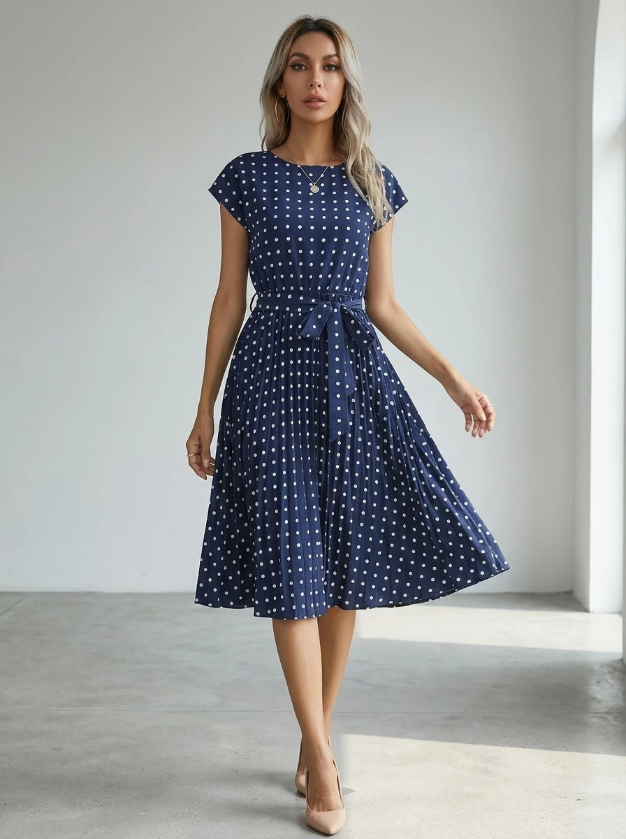 Driftly navy blue polka dot pleated midi dress with cap sleeves and a self-tie waist belt, in a minimalist studio setting with soft window light.
