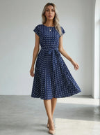 Driftly navy blue polka dot pleated midi dress with cap sleeves and a self-tie waist belt, in a minimalist studio setting with soft window light.