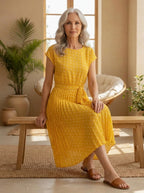 Driftly yellow polka dot pleated midi dress with cap sleeves and a self-tie waist belt, seated on a bench in a light, airy interior space.
