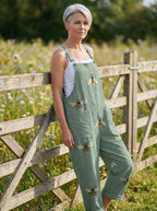 Woman wearing a sage-green bee print sleeveless wide-leg overall jumpsuit leaning against a wooden fence