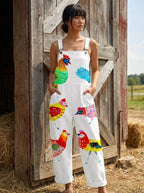 Woman wearing a white colorful rooster and bird print sleeveless wide-leg overall jumpsuit standing near a wooden barn door