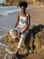 Woman in a cream sardine print jumpsuit sitting on beach rocks
