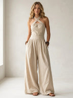 Driftly beige crossed halter wrap jumpsuit with fluid wide-leg trousers and a defined waist, in a minimalist studio setting with soft window light.