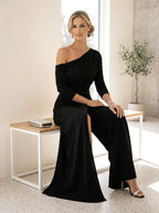 Driftly black asymmetrical one-shoulder jumpsuit with crossed detail and flowing wide-leg trousers, seated on a bench in a light, airy interior space.