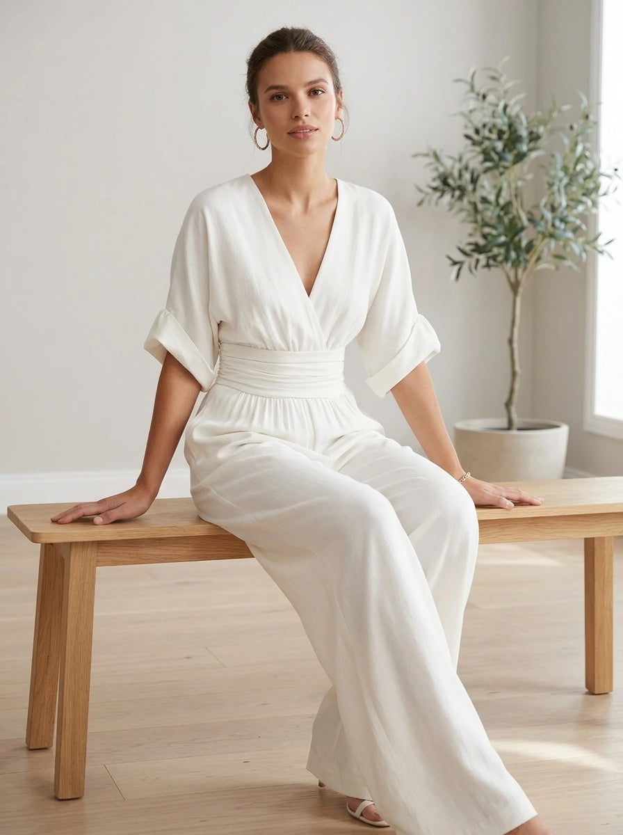 Driftly white deep V-neck jumpsuit with relaxed short sleeves, ruched waist, and fluid wide-leg trousers, seated on a bench in a light, airy interior space.