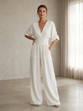 Driftly white deep V-neck jumpsuit with relaxed short sleeves, ruched waist, and fluid wide-leg trousers, in a minimalist studio setting with soft window light.