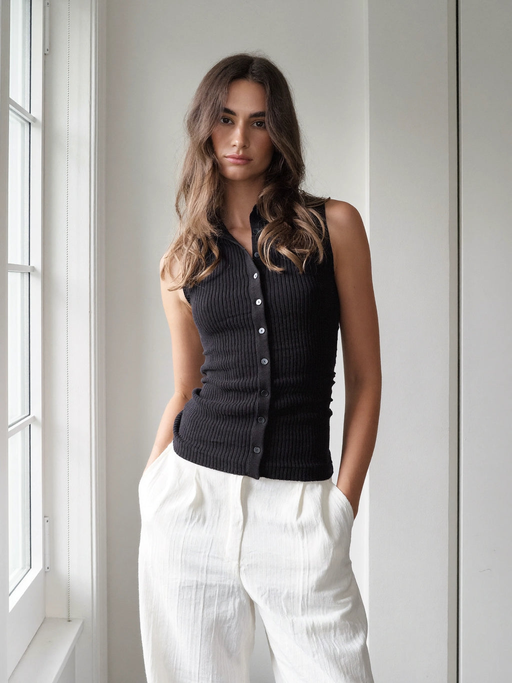 A woman wearing a black ribbed sleeveless button-front vest and white wide-leg pants, standing indoors near a window.