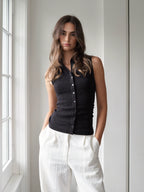A woman wearing a black ribbed sleeveless button-front vest and white wide-leg pants, standing indoors near a window.