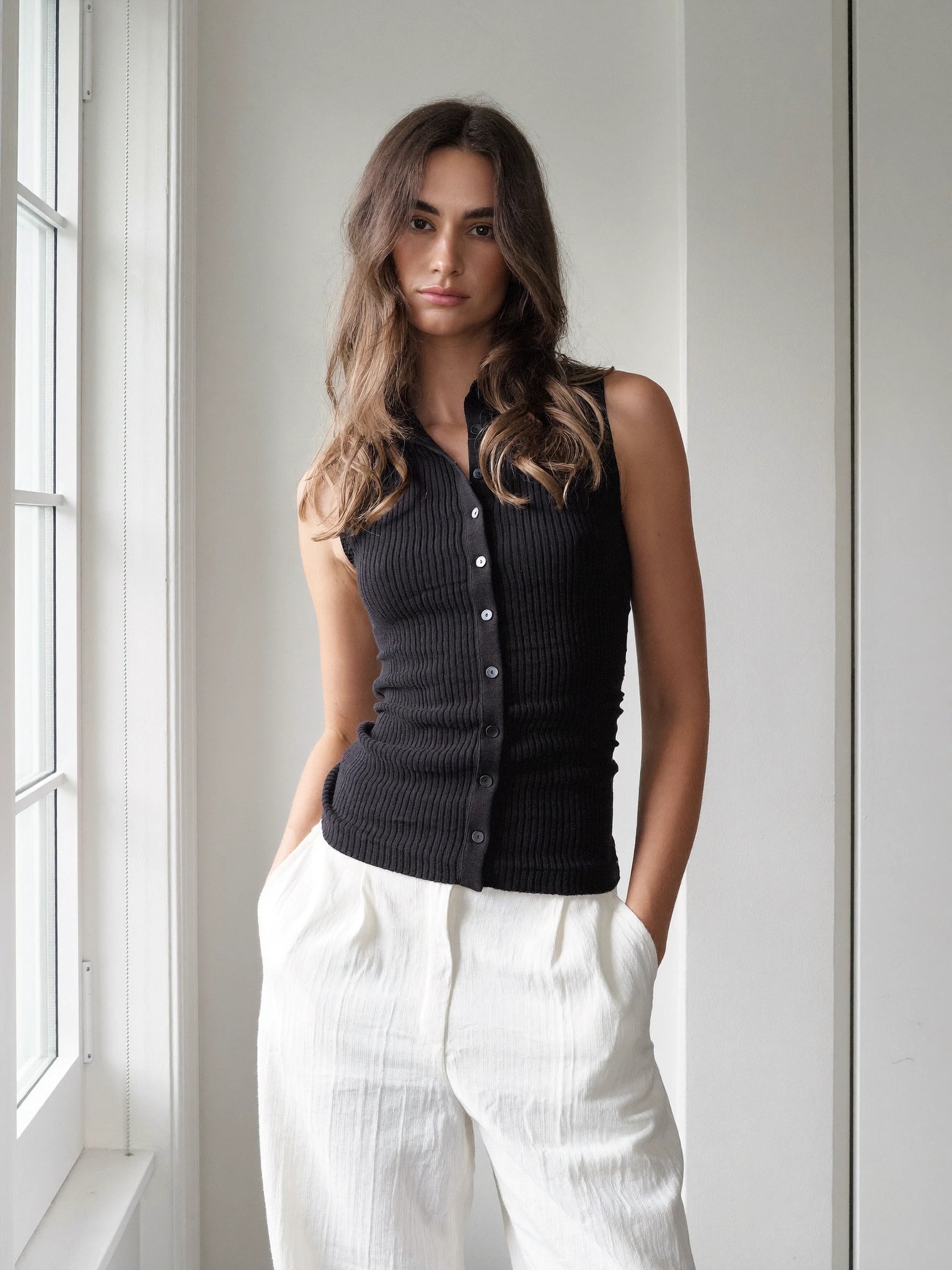 A woman wearing a black ribbed sleeveless button-front vest and white wide-leg pants, standing indoors near a window.