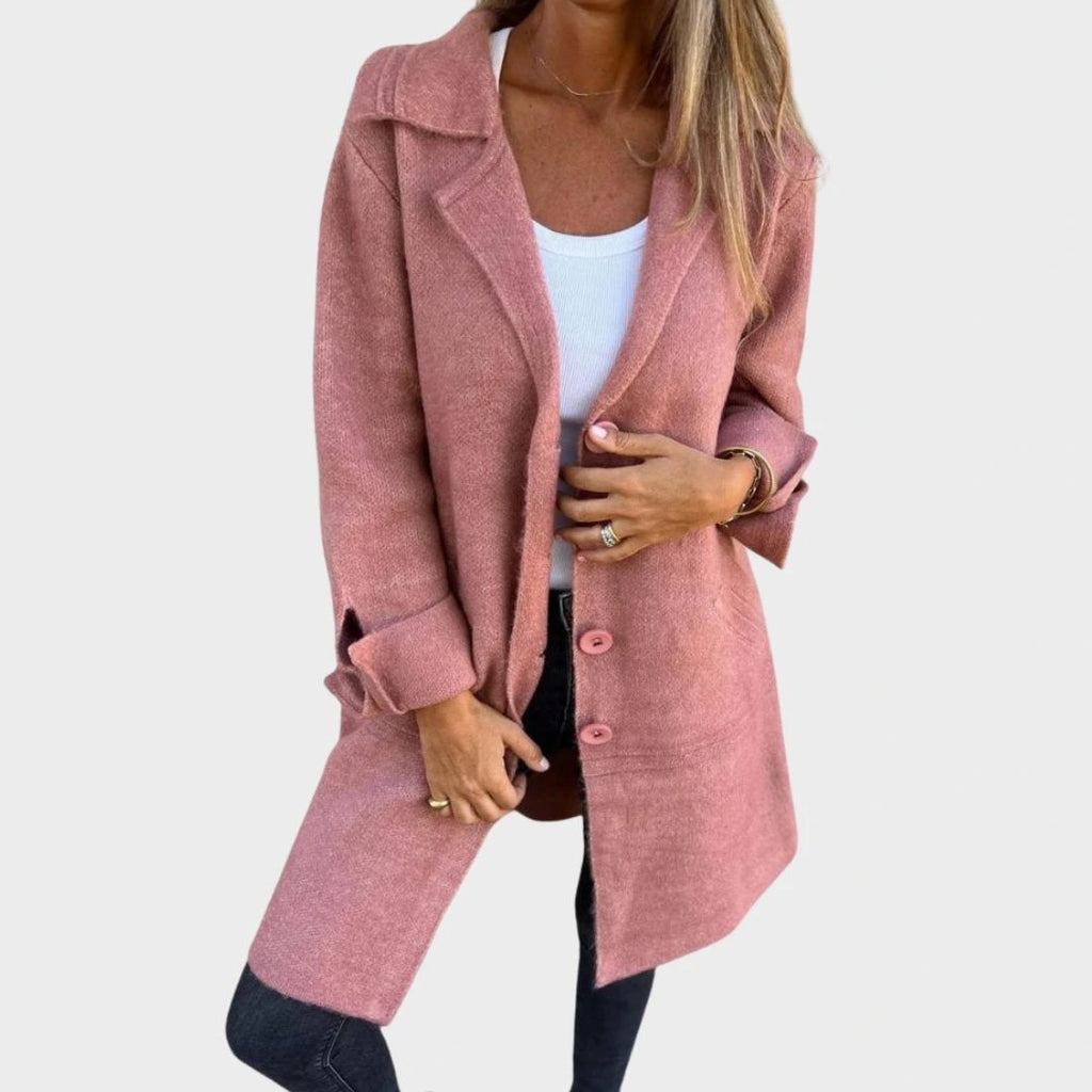 Woman wearing a dusty pink button-up long cardigan coat, alternate pose against a concrete wall