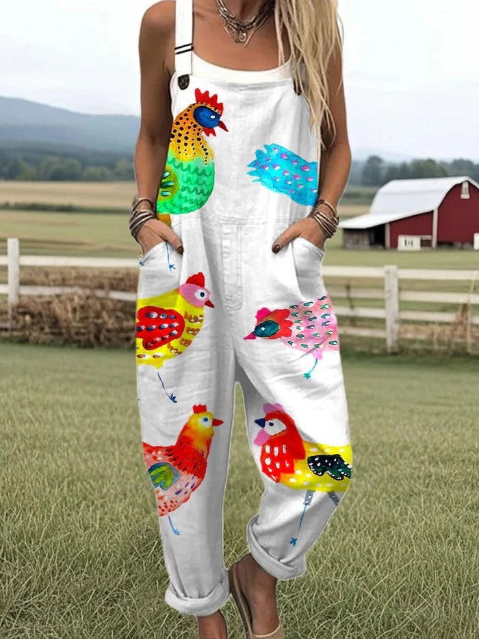 Woman wearing a white colorful rooster and bird print sleeveless wide-leg overall jumpsuit standing in a farm field near a red barn