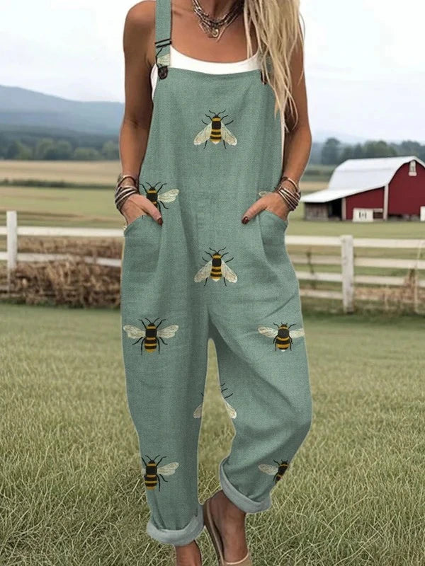 Woman wearing a sage-green bee print sleeveless wide-leg overall jumpsuit standing in a farm field near a red barn