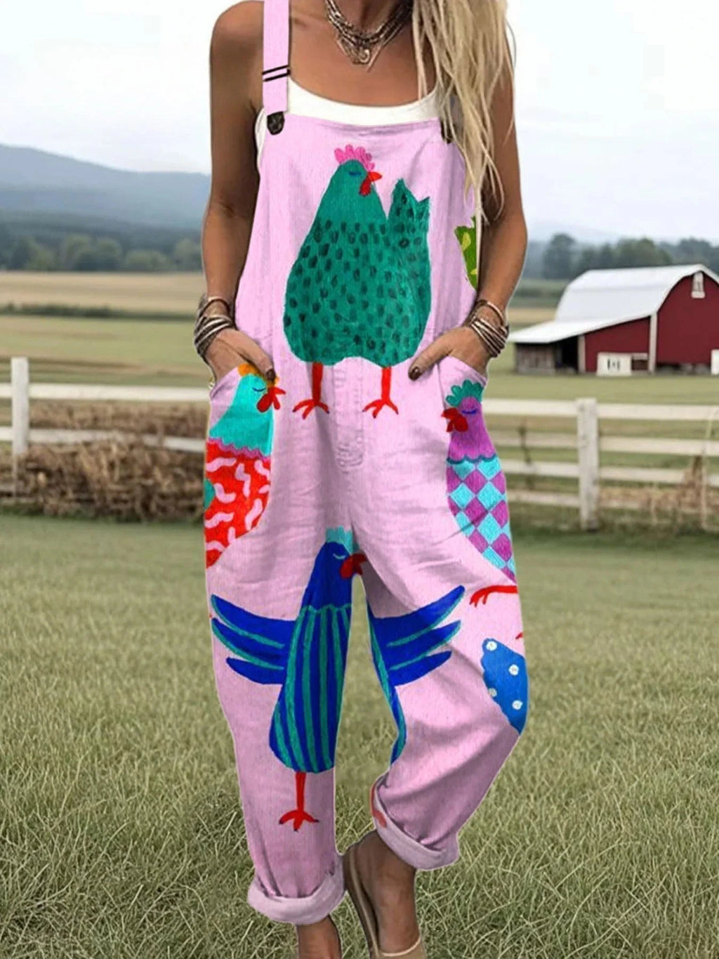 Woman wearing a pink colorful rooster print sleeveless wide-leg overall jumpsuit standing in a farm field near a red barn